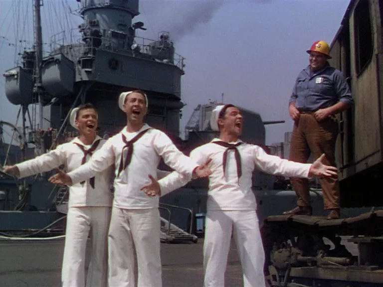 On The Town (1949) movie still: fullbody — A naval base with a large grey warship docked in the background behind sailors. Four men interact on a…