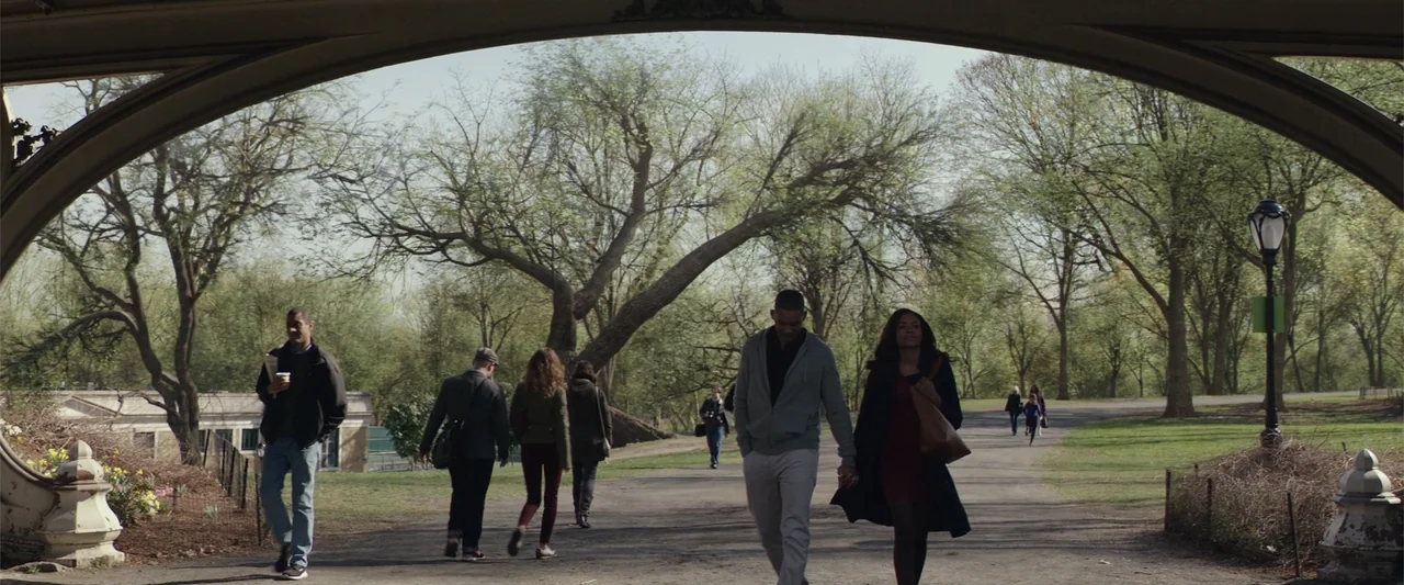 Collateral Beauty (2016) movie still: wide — A stone bridge arch frames a wide view of a tree-lined city park path. A man and woman walk towards the…