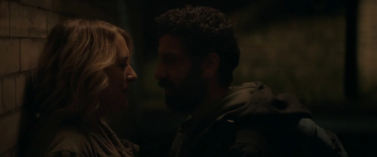 Ali & Ava (2021) movie still: medium — Two individuals stand against a dark brick wall in a dimly lit narrow alleyway. The man faces the blonde…
