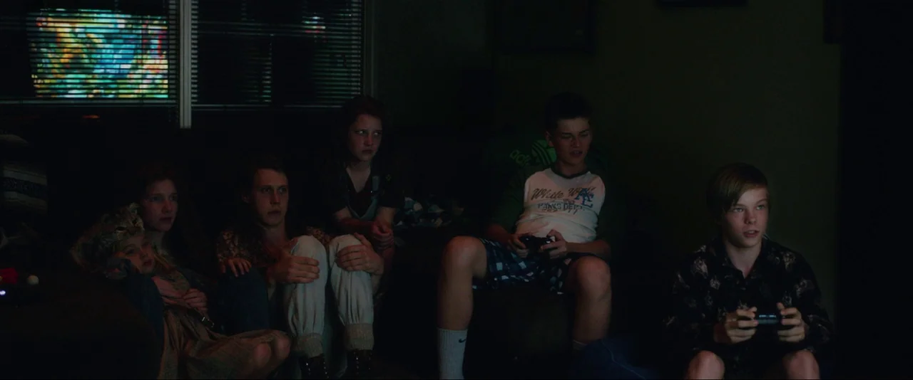 Captain Fantastic (2016) movie still: wide — A dark, cluttered living room contains a couch and multiple seating areas. Six young people are…