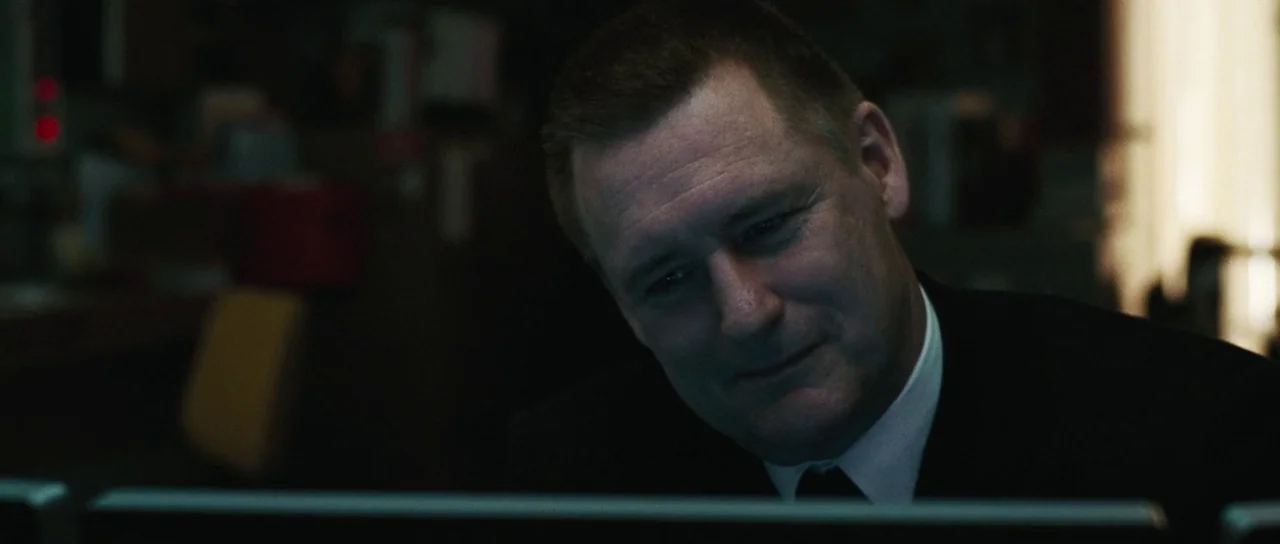 Surveillance (2008) movie still: closeup — A dimly lit office interior at night with a blurred background. A middle-aged man in a suit looks toward…