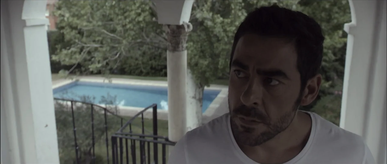 Otro verano (2013) movie still: closeup — A covered terrace with white columns overlooks a rectangular swimming pool in a garden. A man in a white…