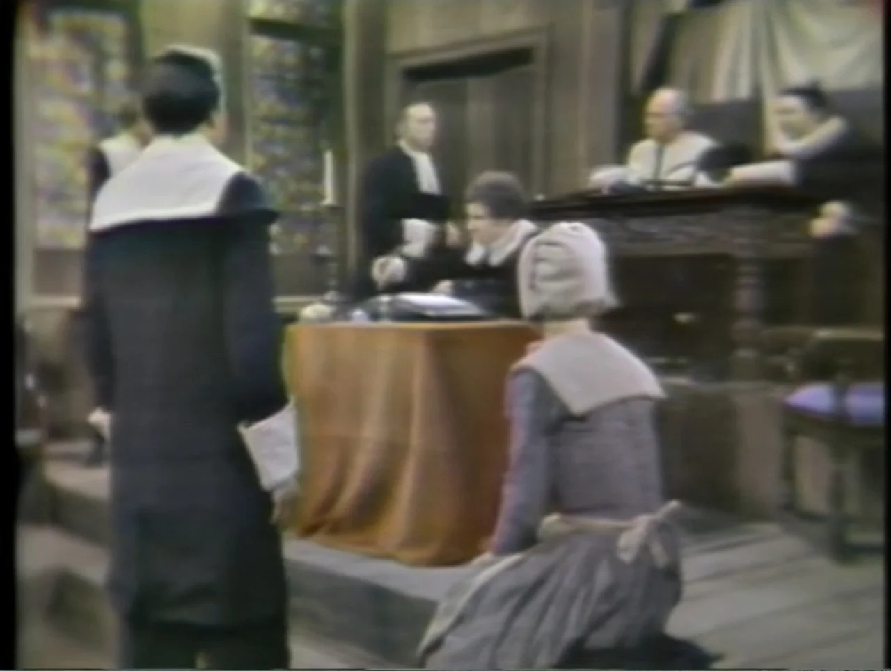 The Crucible (1996) movie still: medium — A historic wooden courtroom setting with formal furniture and period costumes. A woman kneels in the…