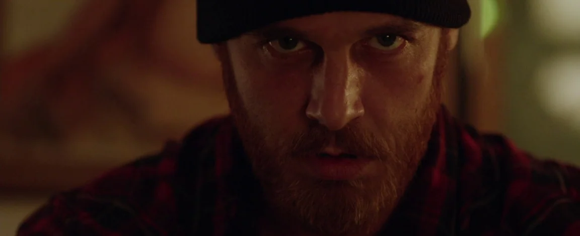 Cheap Thrills (2013) movie still: closeup — A man stands in a dimly lit, dark space with blurred background elements. The man stares directly ahead…