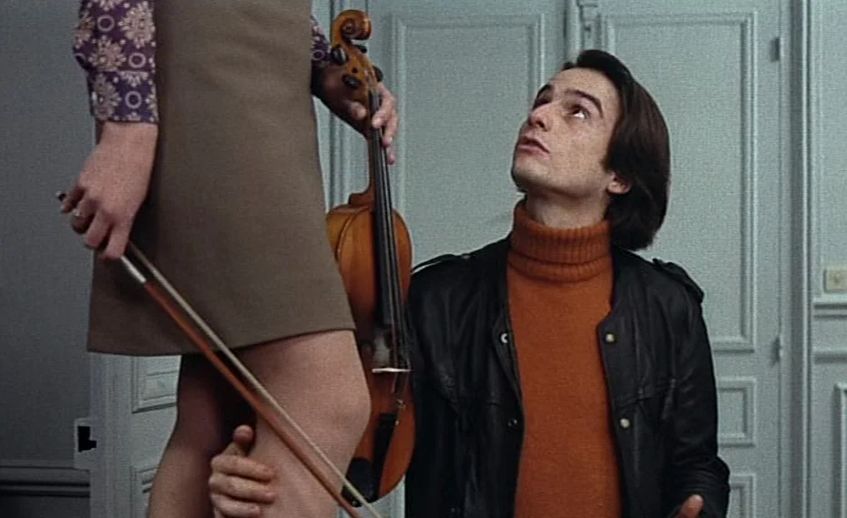 Bed & Board (1970) movie still: medium — A light-walled room with classic French doors in the background. A young man wearing a leather jacket looks…