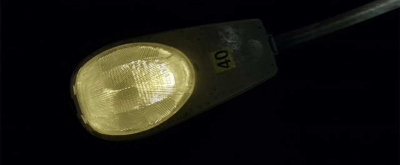 Lights Out (2016) movie still: closeup — A utilitarian street lamp fixture glows brightly against a pitch-black night sky. The metal housing…