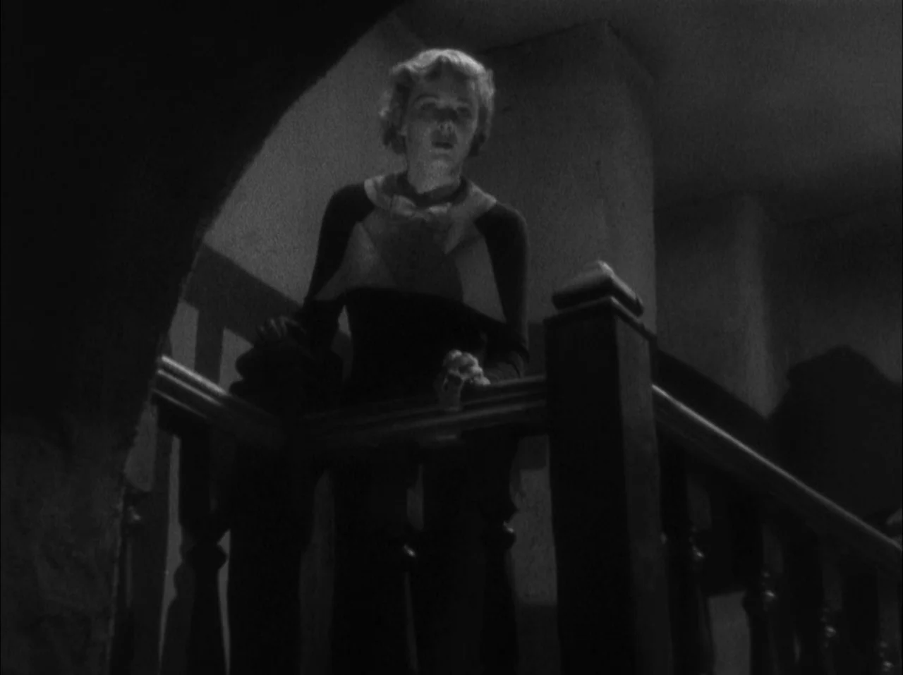 The 39 Steps (1935) movie still: medium — A dark, dimly lit wooden staircase features carved balusters and deep, angular shadows. A woman leans over…
