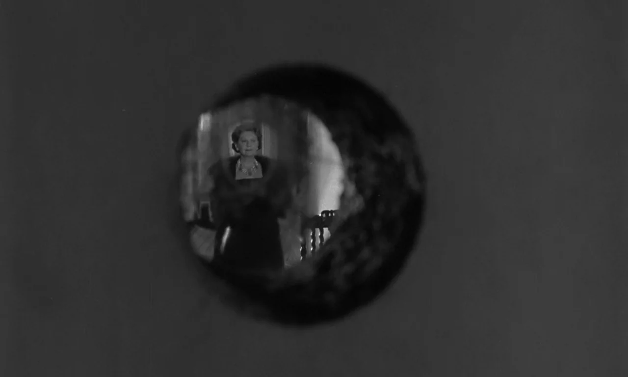 Seance on a Wet Afternoon (1964) movie still: wide — A dimly lit indoor hallway seen from the perspective of a keyhole. The woman stands alone in…