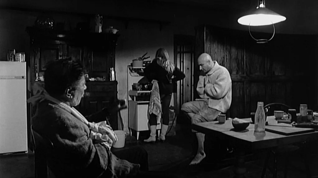 Cul-de-sac (1966) movie still: wide — A dimly lit room features a wooden dining table and vintage cabinetry. Three individuals are gathered in the…