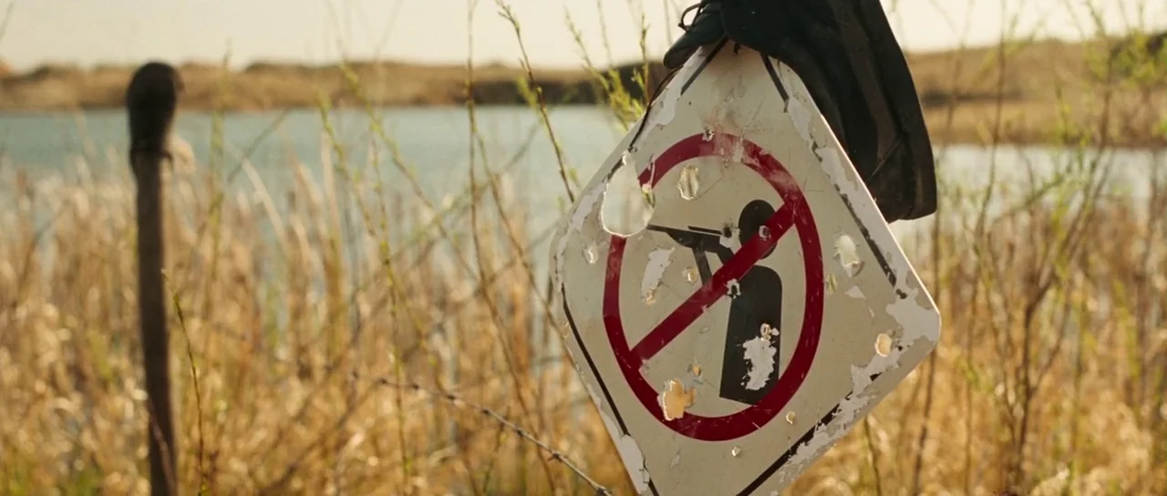 Surveillance (2008) movie still: closeup — A damaged 'no hunting' sign hangs from a wooden post in a grassy marshland. The white metal sign is…