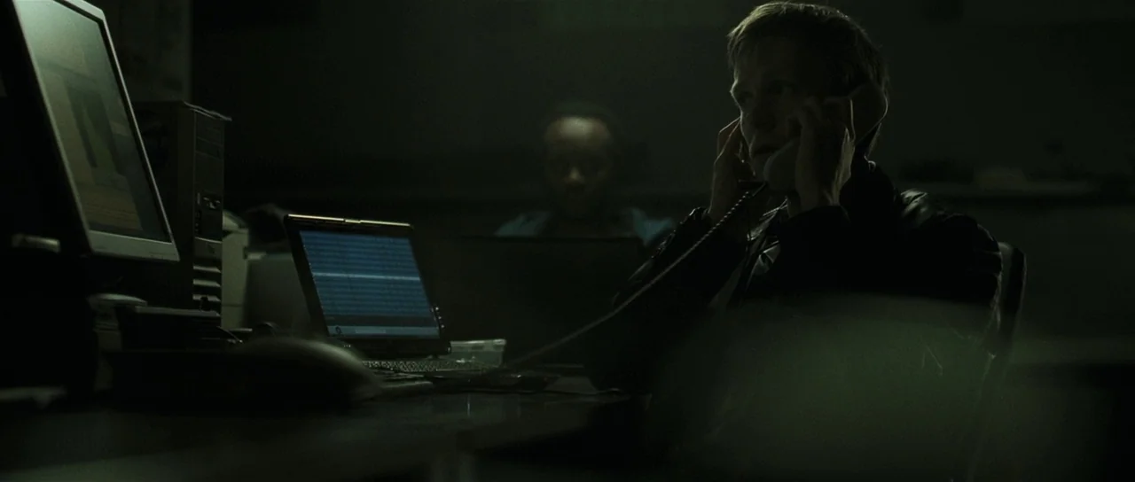 Bullhead (2011) movie still: medium — A dark, cramped office room features multiple computer monitors and cluttered desks. A man in the foreground…