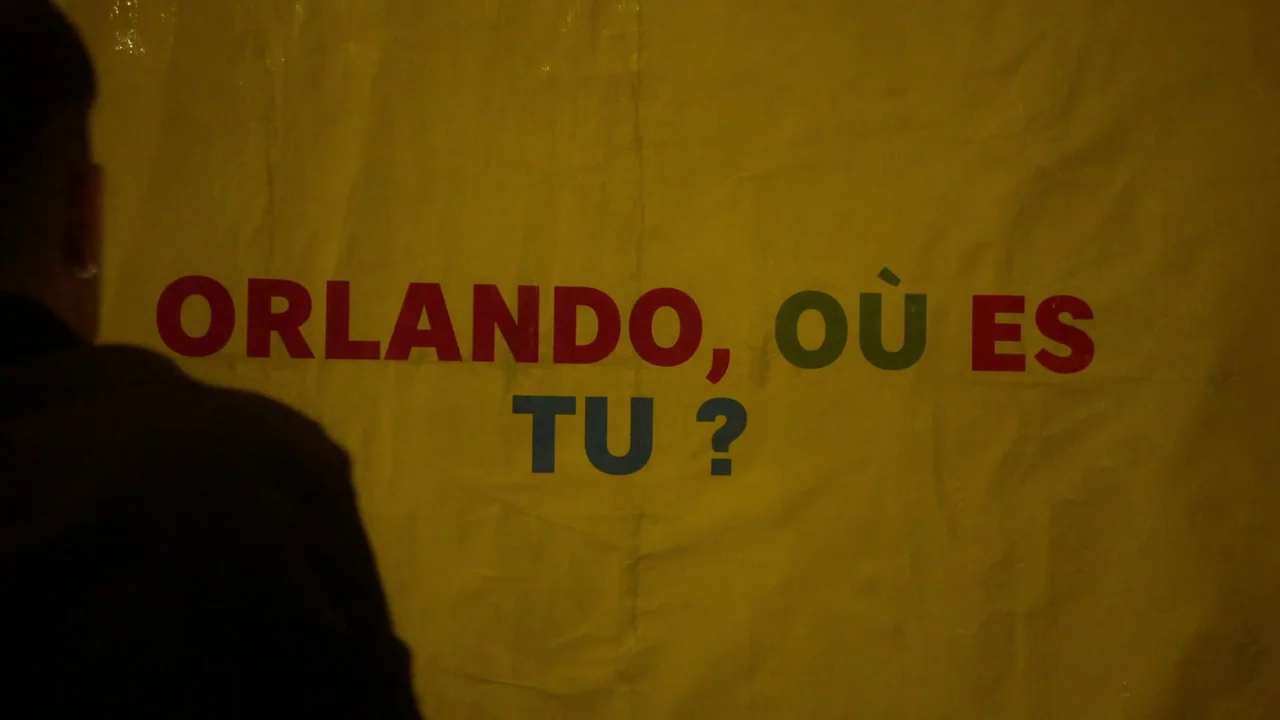 Orlando: My Political Biography (2023) movie still: medium — A person stands before a large yellow fabric banner displayed on a wall. The banner…