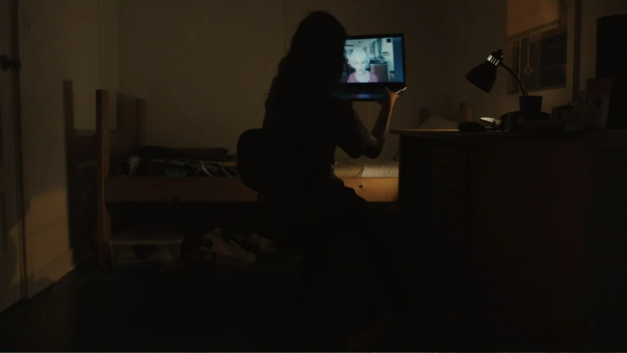 Camp X-Ray (2014) movie still: medium — A dimly lit bedroom features a bed with a wooden frame and a desk. A person sits in silhouette while holding…