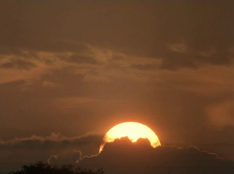 Hail Mary (1985) movie still: establishing — The sun sets behind a dark thick layer of low-hanging clouds. A warm glow illuminates the dense cloud…