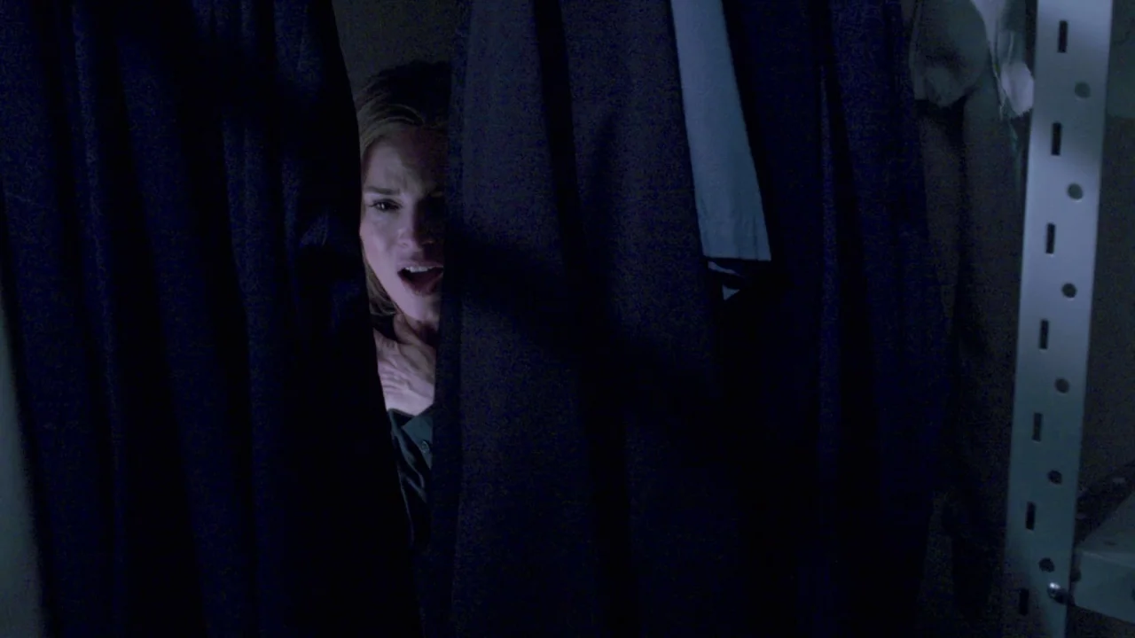 Saw VII: The Final Chapter (2010) movie still: closeup — A dark, cramped space filled with hanging garments on a metal rack. A woman peeks out from…