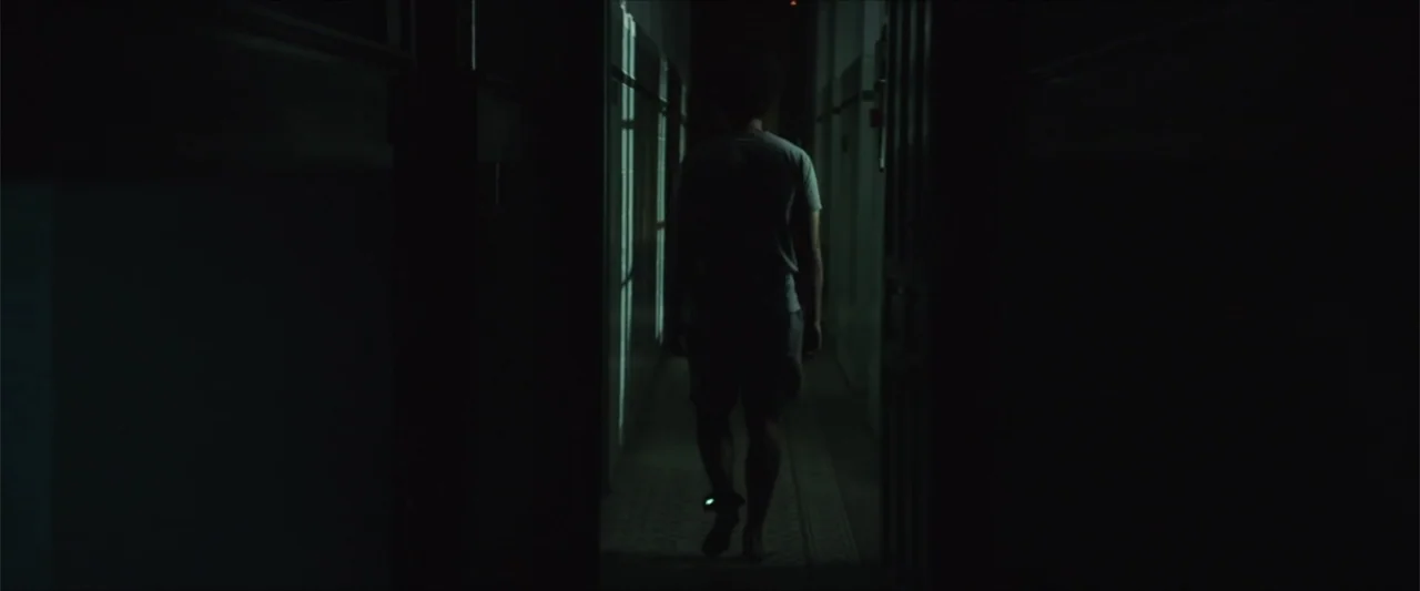 American Carnage (2022) movie still: fullbody — A dark hallway with geometric shadows cast on the tiled floor from a window. A lone person in a…
