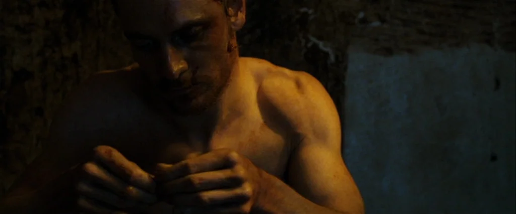 Hunger (2008) movie still: closeup — A dimly lit, bare, industrial-looking space with dark textured stone walls. A shirtless man stares intently at…