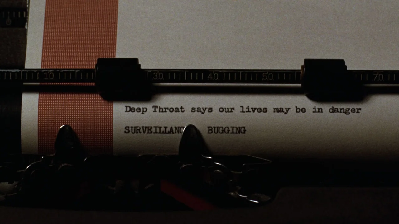All the President's Men (1976) movie still: closeup — A close-up view of a typewriter carriage holding a paper with red-bordered left edge. The text…