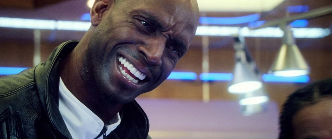 Uncut Gems (2019) movie still: closeup — A man smiles in a dimly lit diner with blue neon lights overhead. He wears a leather jacket and sits near…