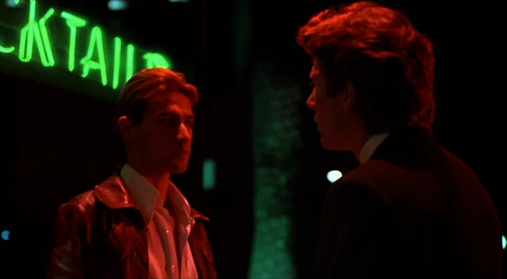 American Gigolo (1980) movie still: medium — A nighttime city sidewalk illuminated by the bright green light of a sign. Two men stand close together…