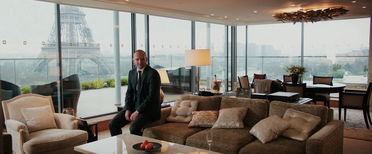 Anna (2019) movie still: fullbody — A luxury suite interior features a panoramic view of the Paris cityscape outdoors. A man in a suit sits perched…
