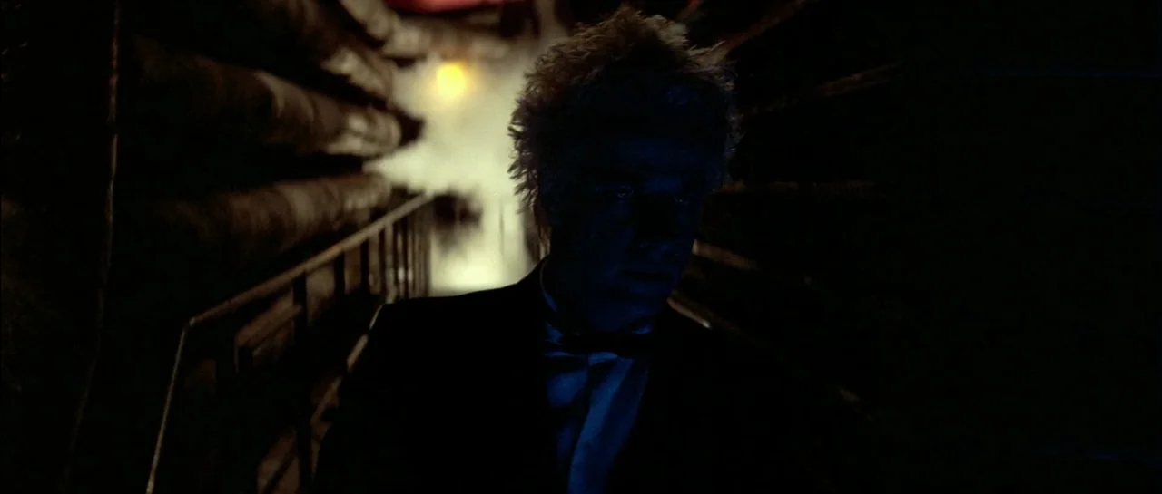 Subway (1985) movie still: medium — A dimly lit narrow industrial corridor contains metal pipes along the ceiling and walls. A man in a suit walks…
