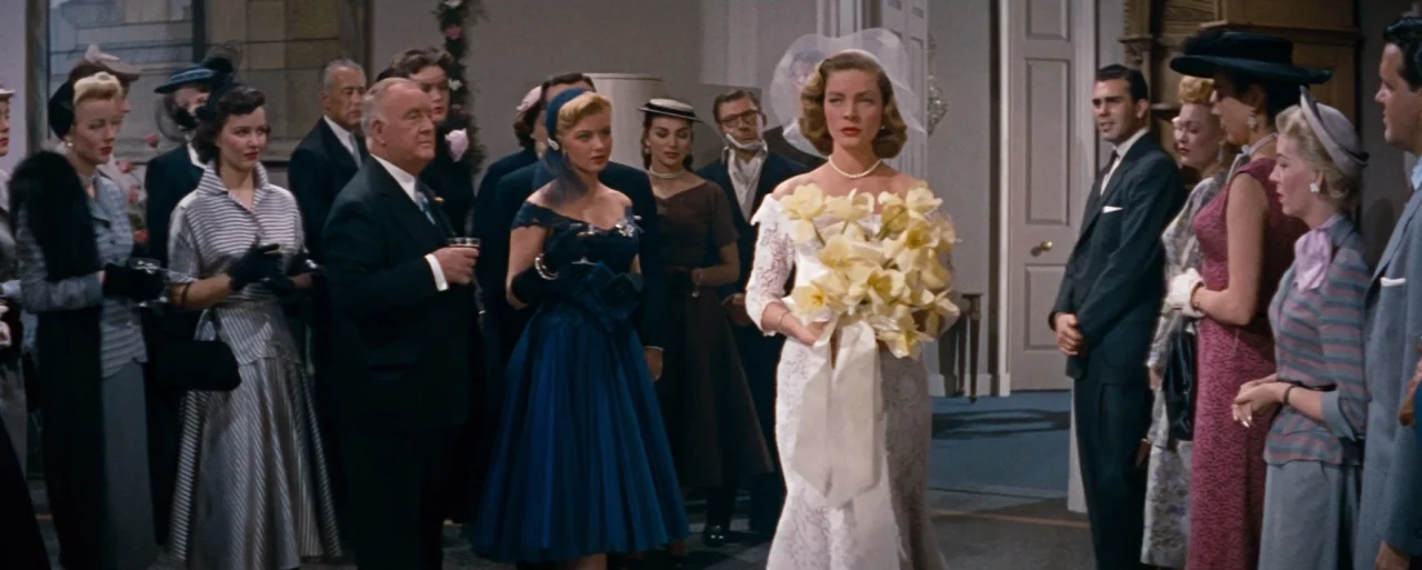 How to Marry a Millionaire movie still: fullbody — A well-lit room is occupied by a group of formally dressed guests. A bride in a white gown and…