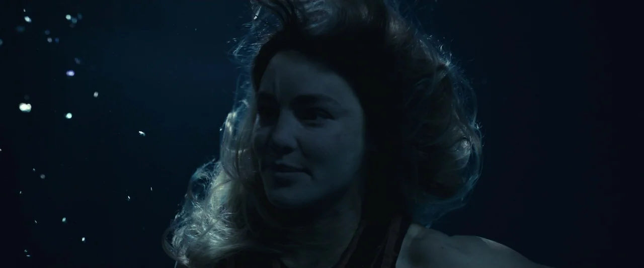 Dangerous Animals (2025) movie still: closeup — A woman submerged in dark deep water with hair drifting upward around her face. She looks forward…