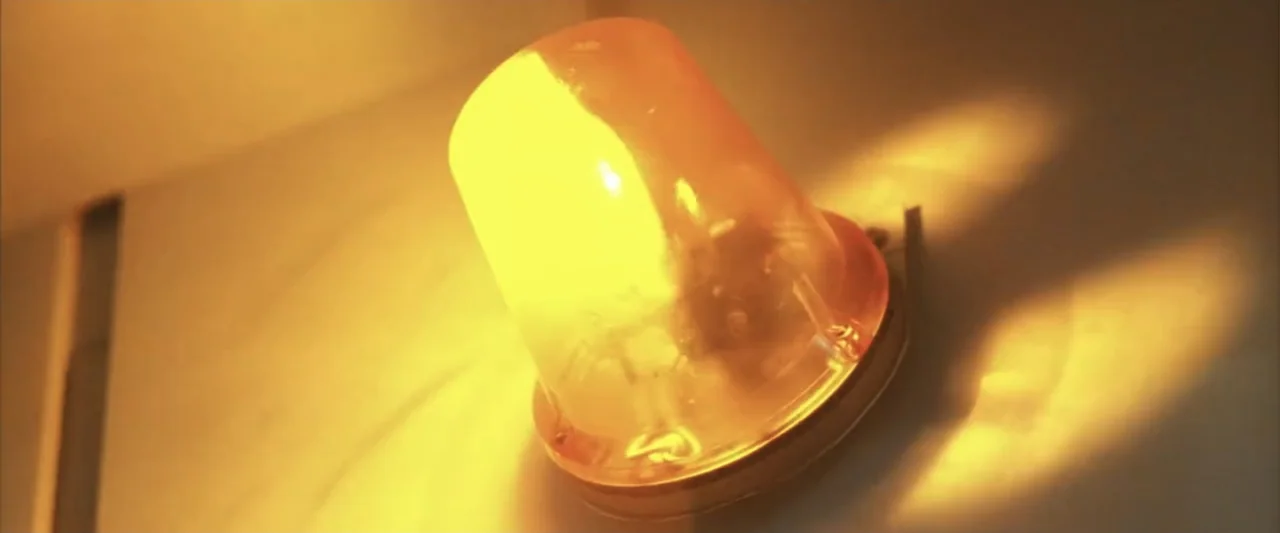A Sound of Thunder (2005) movie still: closeup — A close-up of a bright amber-colored wall-mounted industrial light fixture. The light emits a…