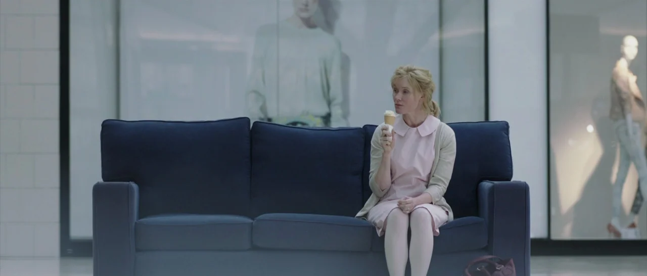 The Babadook (2014) movie still: fullbody — A woman sits alone on a dark blue sofa in a brightly lit shopping mall. She holds an ice cream cone…
