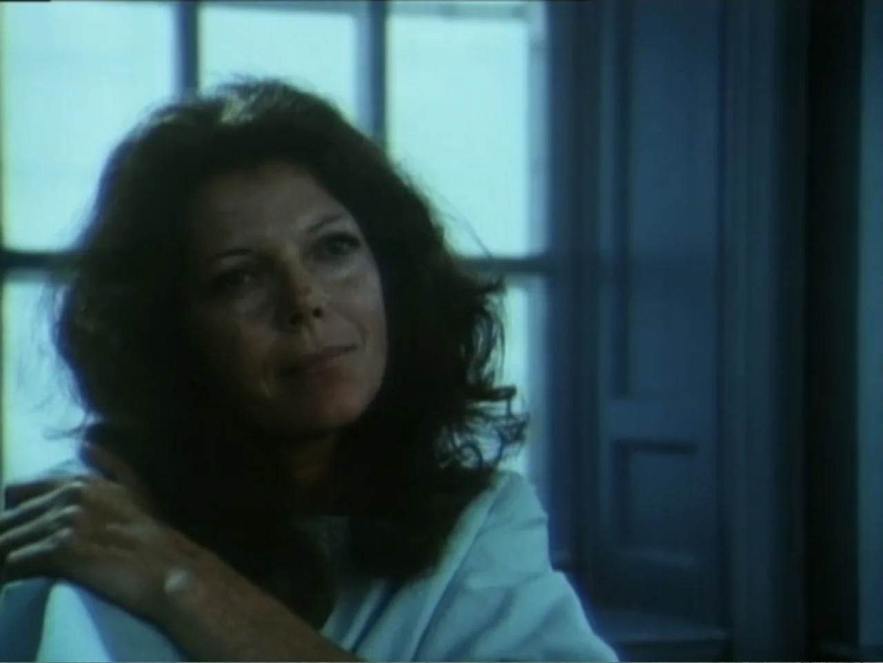Curtains (1995) movie still: medium — A woman sits indoors against a window frame with muted blue tones. She poses with her hand on her shoulder…