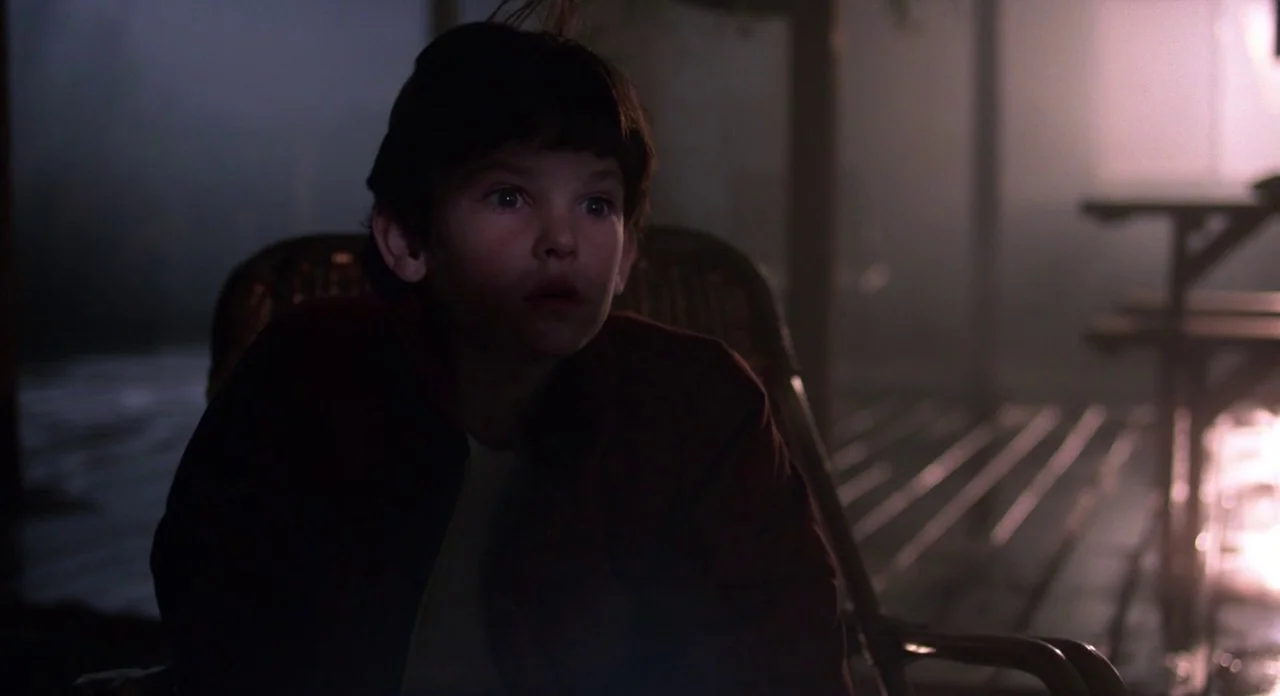 E.T. The Extra-Terrestrial (1982) movie still: medium — A dark wooden porch at night is shrouded in faint mist and low shadows. A young boy sits in…