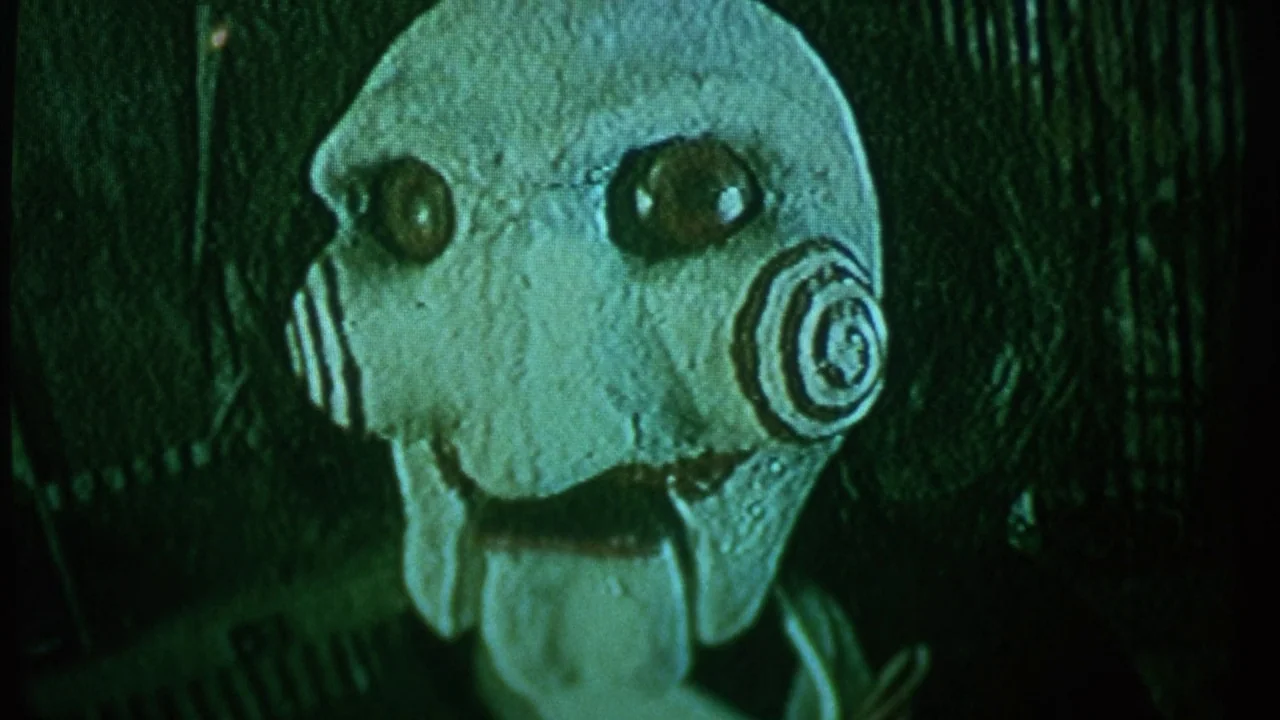 Saw movie still: closeup — The face of a ventriloquist puppet is illuminated by a sharp green-toned light source. The puppet has hollow dark eyes…