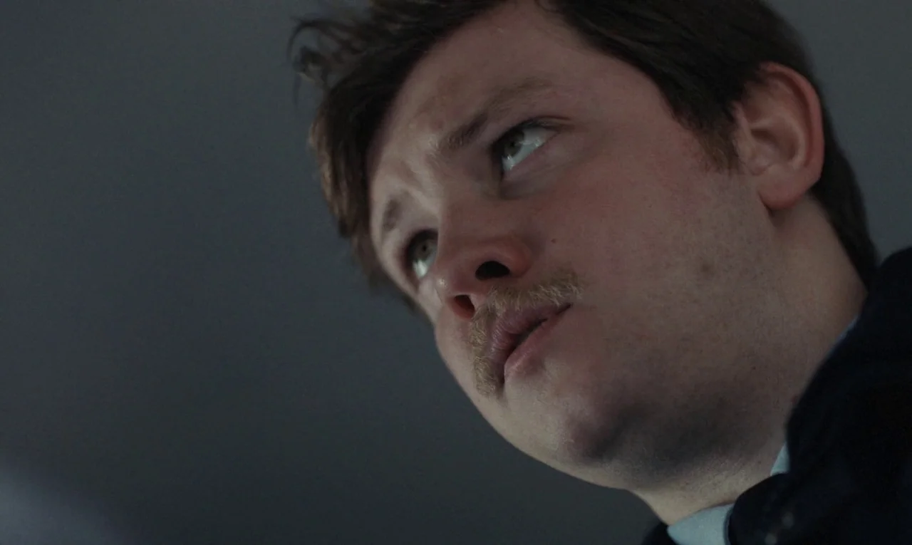 Maldoror (2024) movie still: closeup — A man stands in an underexposed interior space with neutral walls. He tilts his head slightly upward while…