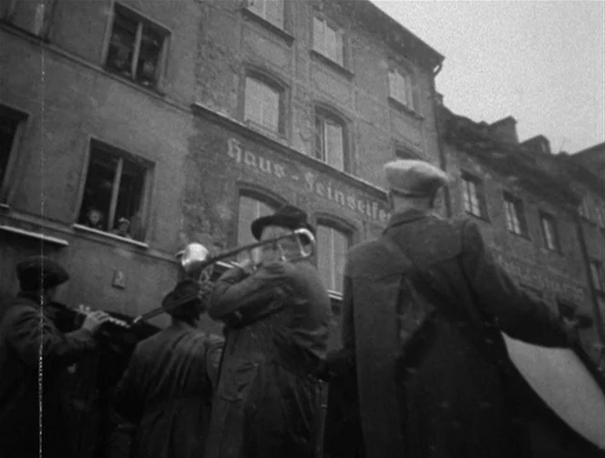 Mr. Arkadin (1955) movie still: medium — A group of street musicians plays in front of an old multi-story building facade. The musicians perform…