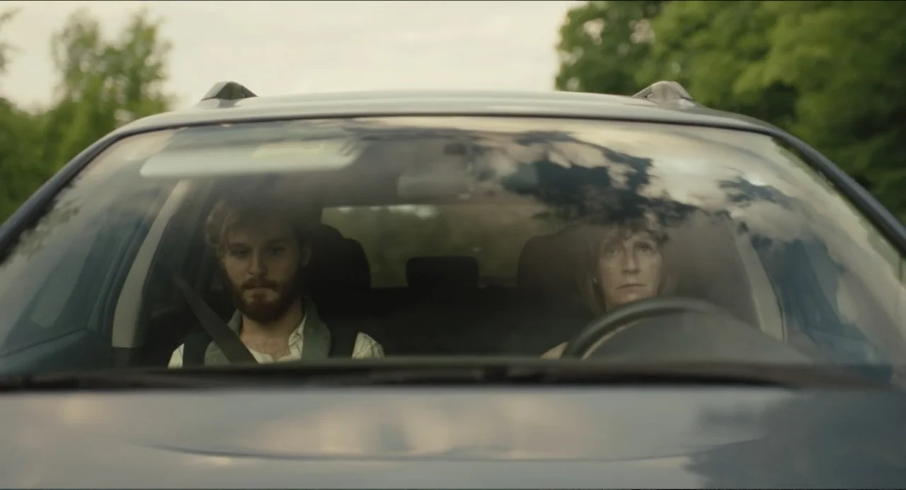 Hell of a Summer (2025) movie still: medium — The interior of a modern vehicle shows two people sitting in the front seats. The man sits in the…