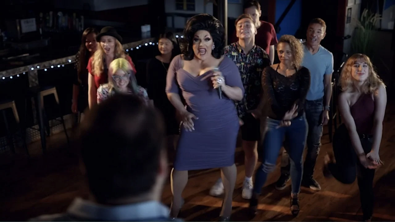 Ask Me to Dance (2022) movie still: fullbody — A dimly lit bar space with a wooden floor and a bar counter behind performers. A person in a purple…