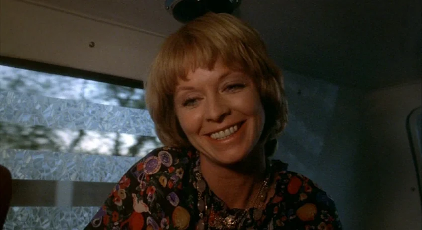 The Silent Partner (1978) movie still: closeup — A woman sits inside a car with patterned window glass beside her. She smiles while looking toward…