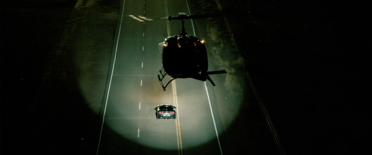 The Last Stand (2013) movie still: wide — A dark asphalt highway is captured at night from a high vantage point above. A small helicopter shines a…