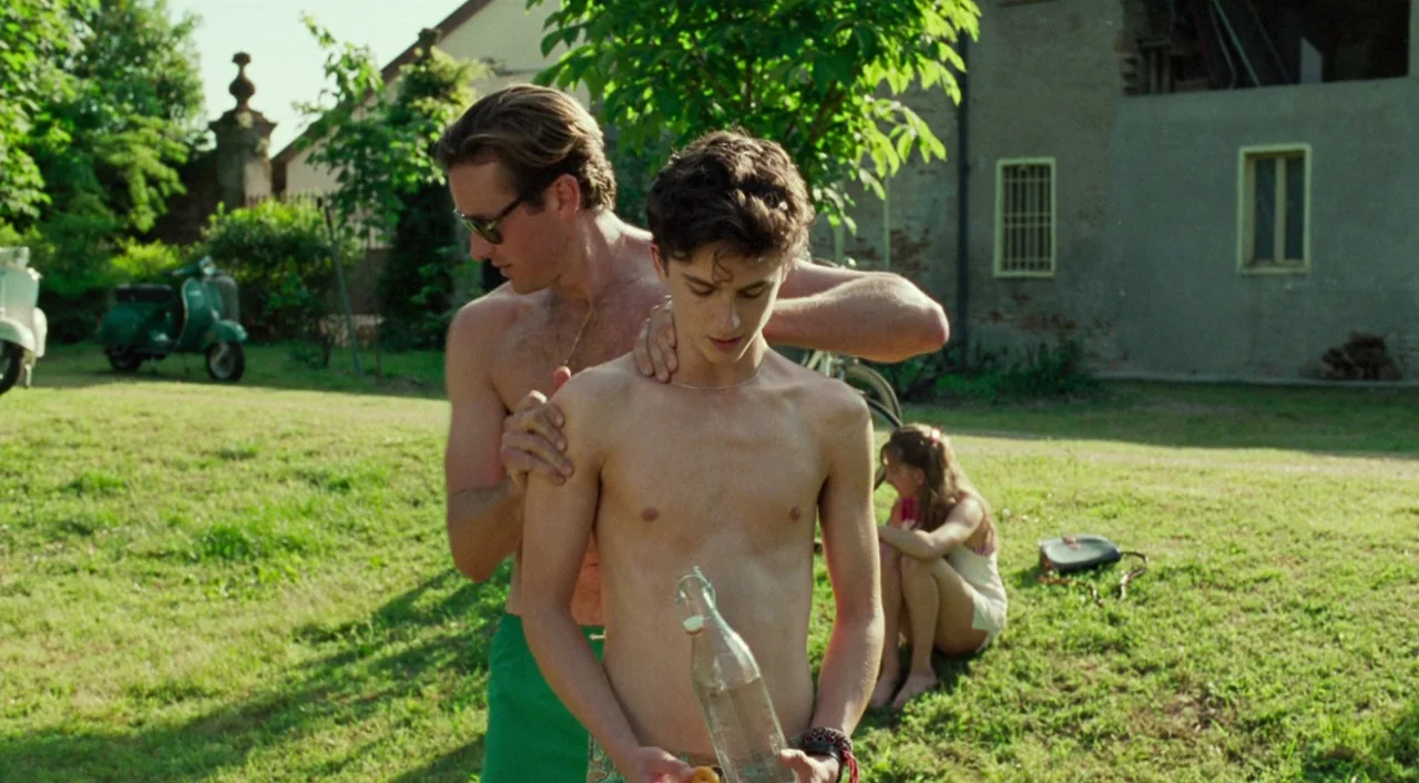 Call Me By Your Name (2017) movie still: medium — A sunny outdoor grassy area with a villa building and parked scooter in background. A shirtless…
