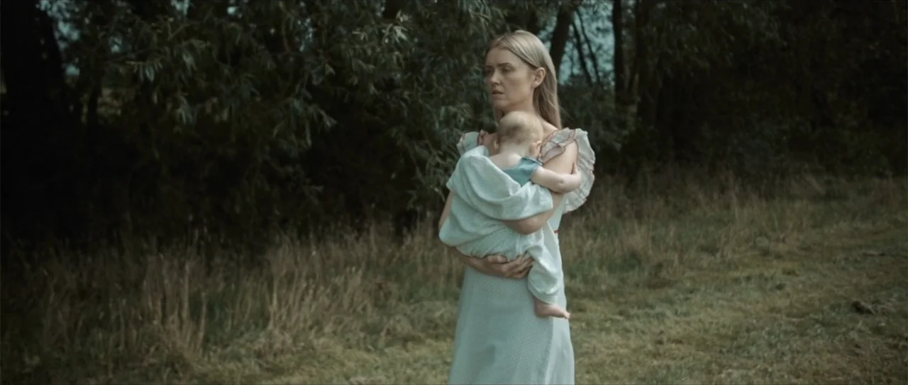 Sin retorno (2009) movie still: medium — A woman in a vintage white dress holds an infant in an overgrown meadow. The background consists of dense…