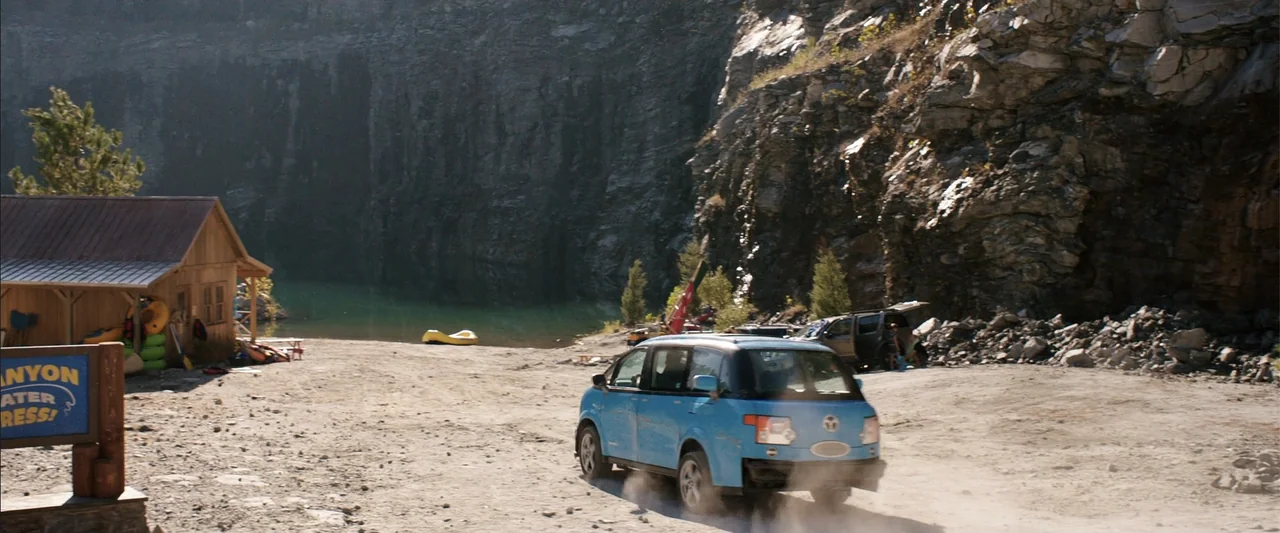 Vacation (2015) movie still: wide — A rugged rock quarry featuring a small wooden building and a calm turquoise pond. A light blue minivan travels…