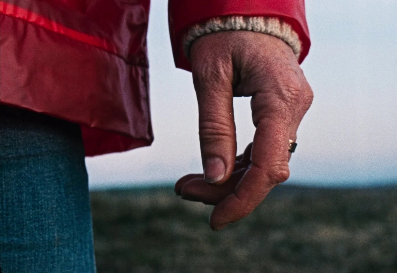 Enys Men (2022) movie still: closeup — A person stands in an open landscape wearing a red waterproof coat. Their hand hangs down at their side…