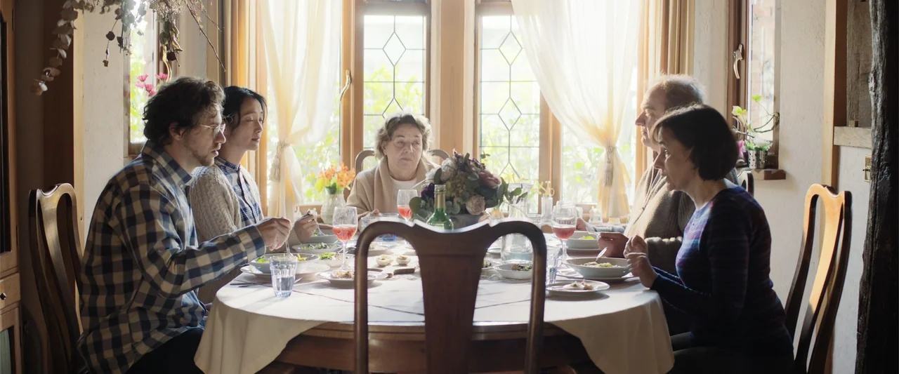 Angel Flight THE MOVIE (2026) movie still: medium — A sunlit dining room features a round table surrounded by wooden chairs and window. Five adults…