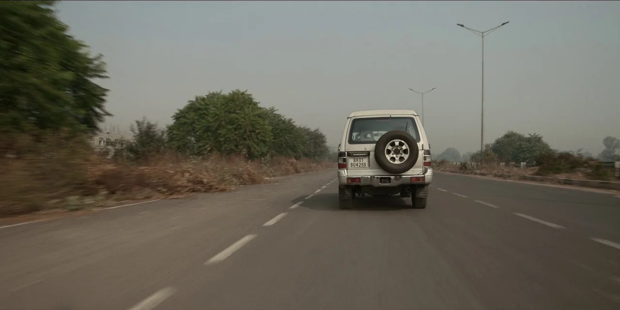 The White Tiger (2021) movie still: wide — An asphalt highway stretches into the distance under a pale, hazy daytime sky. A silver SUV drives away…