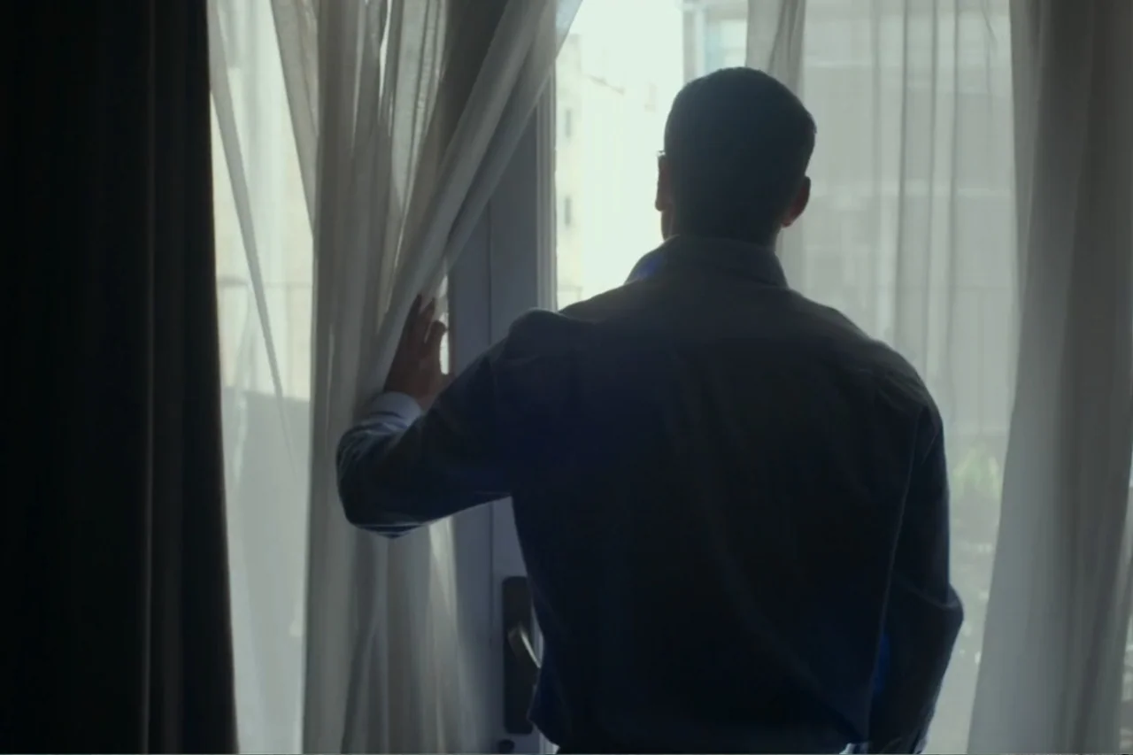 Cross the Line (2020) movie still: medium — A modern room with bright sunlight streaming through a large window covered by curtains. A man in a blue…