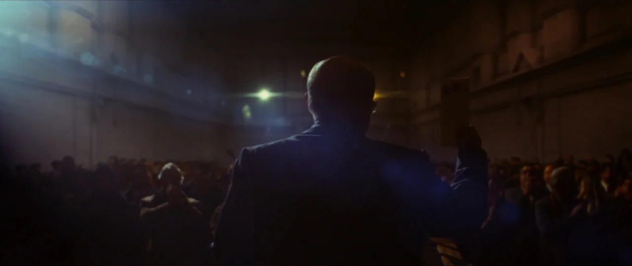 Neruda (2016) movie still: medium — A dimly lit, large interior hall with rows of seated, blurred attendees in shadow. A man stands at a podium in…