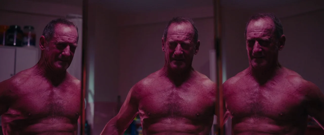 Titane (2021) movie still: medium — A dimly lit room with multiple mirrors displaying the same subject from different angles. An athletic older man…