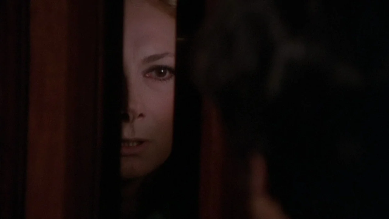 The Black Belly of the Tarantula (1971) movie still: closeup — A woman peeks through a narrow vertical gap in a dark wooden door. Only half of her…