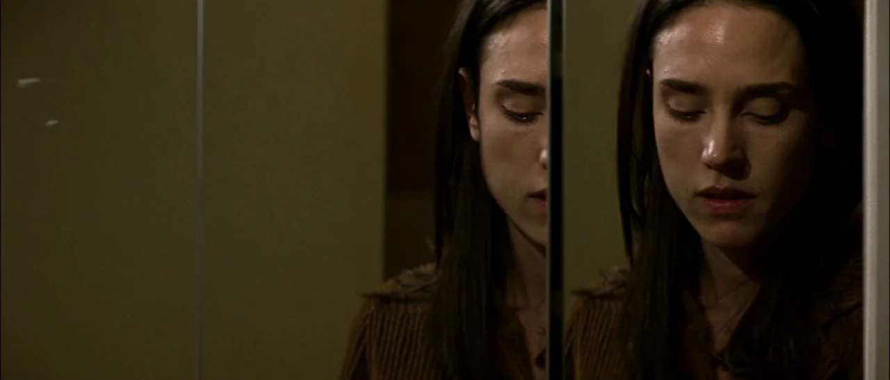 Dark Water (2005) movie still: closeup — A close view of a woman looking at her reflection in a medicine cabinet. The mirror splits her face into…